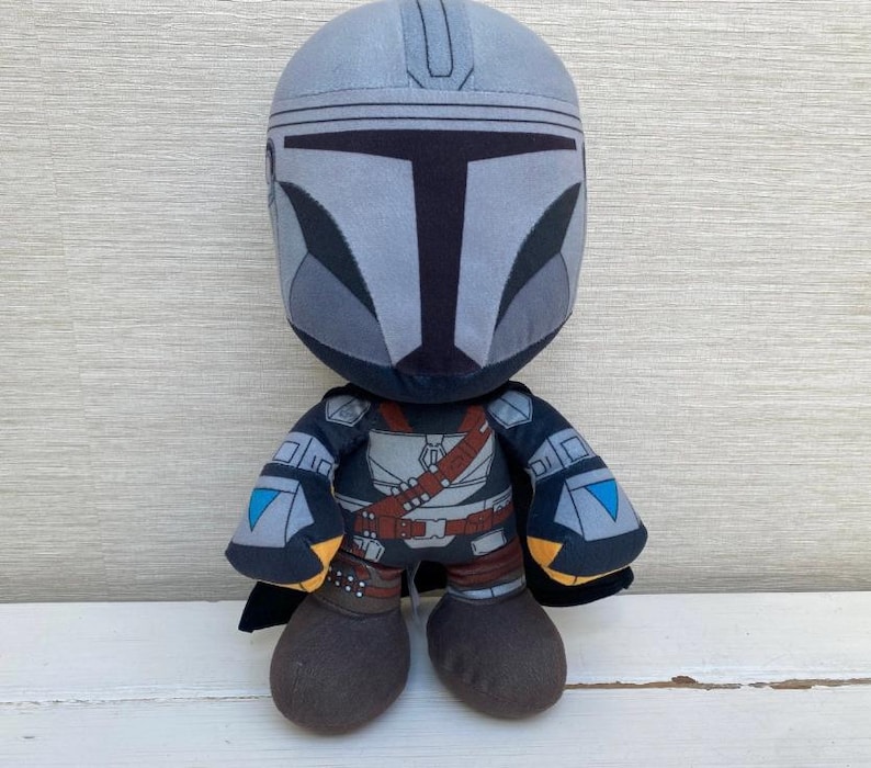 May include: A plush toy depicting a character from a popular science fiction series. The figure has a gray helmet and a detailed outfit with brown and black accents. The toy has a black cape and brown boots.