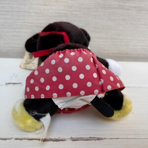 K&ouml;nnte beinhalten: Eine Minnie Mouse Pl&uuml;schpuppe, von hinten gesehen, mit schwarzem K&ouml;rper, roter Schleife und rotem Rock mit wei&szlig;en Punkten. Die Puppe hat gelbe F&uuml;&szlig;e und einen wei&szlig;en Schwanz. Ein Etikett ist angebracht.