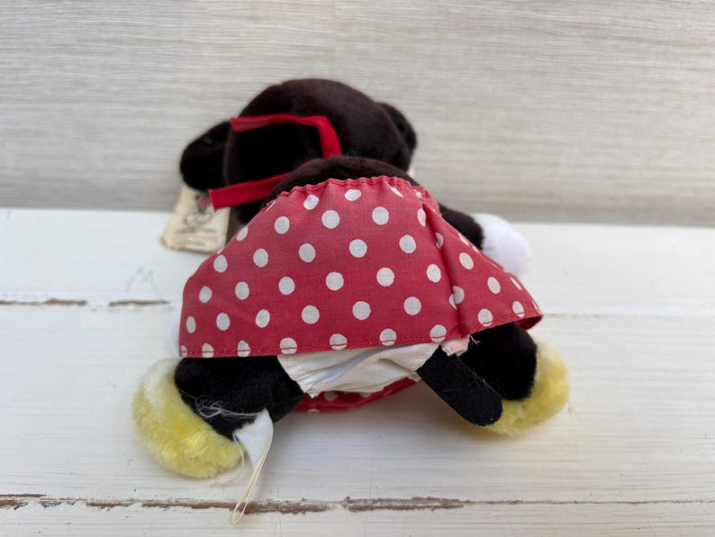 K&ouml;nnte beinhalten: Eine Minnie Mouse Pl&uuml;schpuppe, von hinten gesehen, mit schwarzem K&ouml;rper, roter Schleife und rotem Rock mit wei&szlig;en Punkten. Die Puppe hat gelbe F&uuml;&szlig;e und einen wei&szlig;en Schwanz. Ein Etikett ist angebracht.