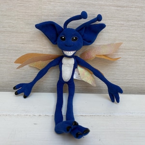 May include: A plush toy creature with deep blue fabric, large pointed ears, and two antennae. It features a wide grin with visible white teeth, a light-colored belly patch, and iridescent, translucent wings extending from its back.