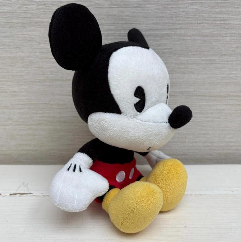 Op de afbeelding: Een pluche Mickey Mouse-speelgoed, met een zwart-witte kop, grote oren, een zwarte neus en een rood shirt met witte knopen. Het speelgoed heeft witte handschoenen en gele voeten en zit.