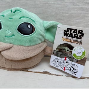 May include: A Grogu plush toy from The Mandalorian series. The doll features large black eyes, a light green face and ears, and a tan robe. A Star Wars Mandalorian tag is attached, with the text "Recycled Made With Stuffing."