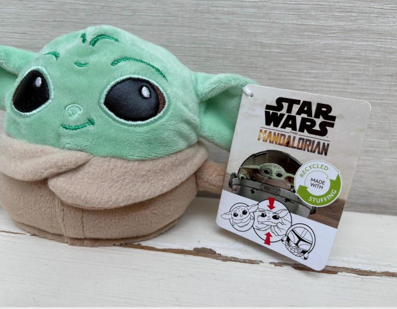 May include: A Grogu plush toy from The Mandalorian series. The doll features large black eyes, a light green face and ears, and a tan robe. A Star Wars Mandalorian tag is attached, with the text "Recycled Made With Stuffing."