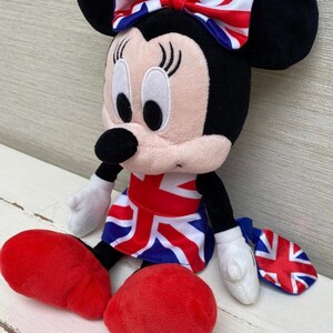 Puede incluir: Un peluche de Minnie Mouse con un lazo, un vestido y un accesorio con el dise&ntilde;o de la Union Jack. La mu&ntilde;eca tiene orejas negras, cara rosa y zapatos rojos. La mu&ntilde;eca est&aacute; sentada sobre una superficie blanca.