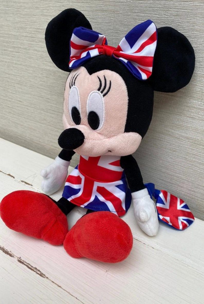 Puede incluir: Un peluche de Minnie Mouse con un lazo, un vestido y un accesorio con el dise&ntilde;o de la Union Jack. La mu&ntilde;eca tiene orejas negras, cara rosa y zapatos rojos. La mu&ntilde;eca est&aacute; sentada sobre una superficie blanca.