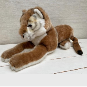 Peut inclure: Une peluche de couguar en position détendue. Le couguar a une fourrure brune avec des accents blancs et beiges sur le visage, la poitrine et les pattes. La queue a une pointe noire. Le jouet est posé sur une surface blanche.