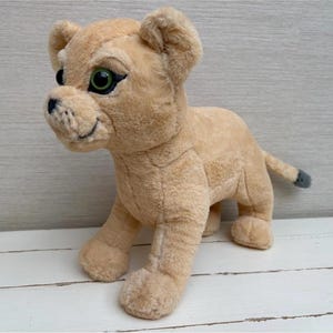 May include: A soft toy lion cub in a standing position. The cub features tan fur, green eyes, and a black nose. The tail has a grey tip. The toy is made of a soft material and is designed to resemble a lion cub.
