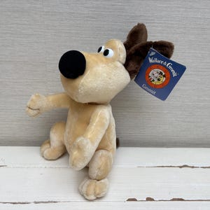 Puede incluir: Un juguete de peluche de perro color canela con una nariz negra grande, ojos blancos y orejas de color marrón oscuro. El animal de peluche tiene una etiqueta de cartón azul unida a la oreja con el texto Wallace & Gromit y Gromit en letras blancas y amarillas.