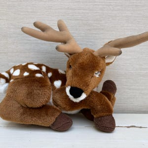 May include: A soft toy deer with brown fur, white spots, and tan antlers. The deer has a black nose, brown hooves, and is in a lying position. The toy is designed to look like a fawn.