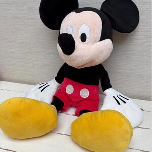 May include: A plush Mickey Mouse doll with black ears, a black body, red shorts, and yellow shoes. The doll has white gloves and a smiling face. The toy is sitting on a white wooden surface.