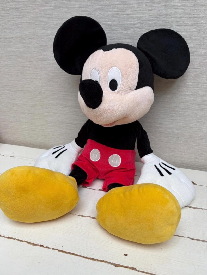 May include: A plush Mickey Mouse doll with black ears, a black body, red shorts, and yellow shoes. The doll has white gloves and a smiling face. The toy is sitting on a white wooden surface.