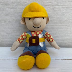 Op de afbeelding: Een pluche pop van een bouwvakker met een felgele veiligheidshelm, een rood-geel geruit shirt met lange mouwen, een blauwe tuinbroek in spijkerstofstijl en een bruine gereedschapsriem met zilverkleurige accenten. De pop heeft gele laarzen.