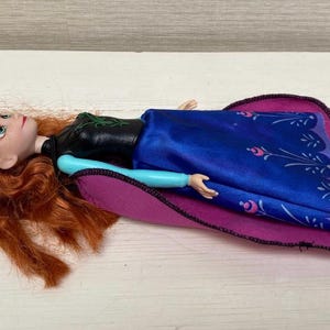 May include: A doll with red hair, dressed in a black top, blue dress, and black boots, lying on a purple and pink blanket. The doll's arms are light blue. A children's toy.