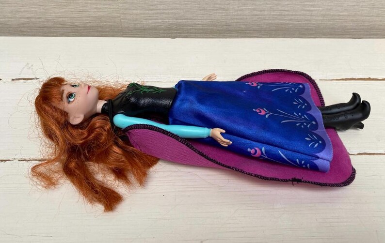 May include: A doll with red hair, dressed in a black top, blue dress, and black boots, lying on a purple and pink blanket. The doll's arms are light blue. A children's toy.