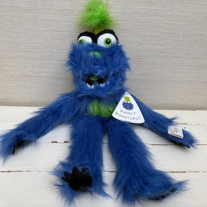 Peut inclure: Une marionnette monstre bleue avec des détails verts et une crête verte. La marionnette a de grands yeux globuleux et un sourire édenté. Une étiquette indique "Puppet Monsters!!!". La marionnette est conçue pour être utilisée comme marionnette à main.