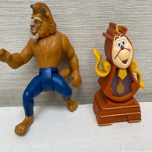 Può includere: Due statuette di plastica del film d'animazione La Bella e la Bestia. Uno è un personaggio della Bestia marrone e beige con pantaloni blu. L'altro è un personaggio dell'orologio Cogsworth marrone e beige.