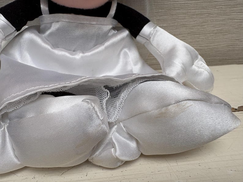 May include: A plush toy of a character wearing a white satin wedding dress. The dress has a layered skirt and spaghetti straps. The character's arms and gloves are white, and the head has black ears and a black bow.