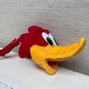 May include: A plush Woody Woodpecker keychain. The red plush bird has a yellow beak and a red clip. The eyes are white with black pupils and green irises. The keychain is on a white surface.