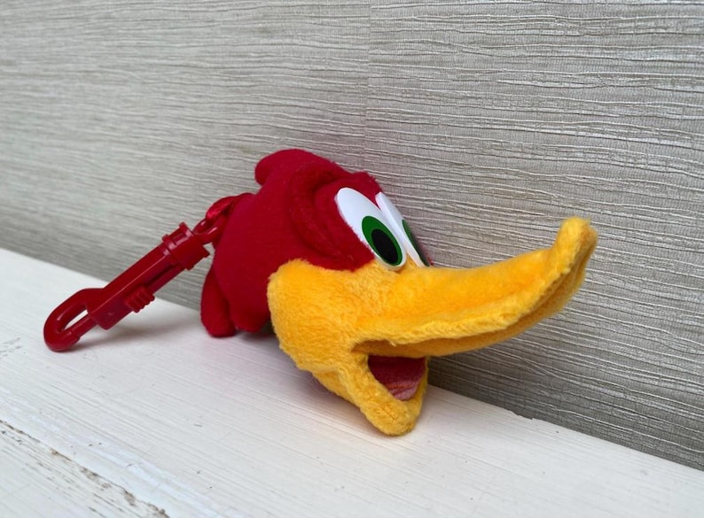 May include: A plush Woody Woodpecker keychain. The red plush bird has a yellow beak and a red clip. The eyes are white with black pupils and green irises. The keychain is on a white surface.