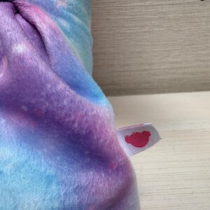 May include: A plush, soft fabric in shades of blue, purple, and pink, resembling a galaxy. A small white tag with a pink heart-shaped logo is attached to the fabric. The fabric is folded, showing the texture and color variations.