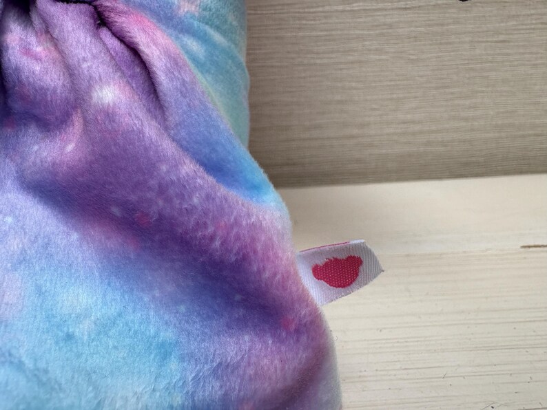 May include: A plush, soft fabric in shades of blue, purple, and pink, resembling a galaxy. A small white tag with a pink heart-shaped logo is attached to the fabric. The fabric is folded, showing the texture and color variations.