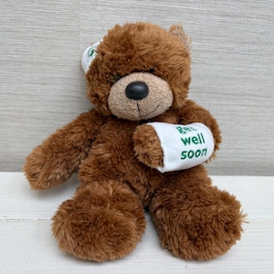 Könnte beinhalten: Ein brauner Teddybär mit schwarzer Nase und einem weißen Verband am Arm mit der Aufschrift "Get well soon". Der Bär hat auch einen weißen Verband am Kopf. Der Bär sitzt auf einer weißen Oberfläche.