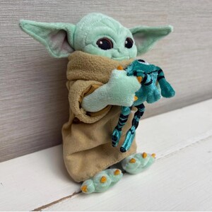 May include: A plush Grogu doll, also known as Baby Yoda, in a tan robe, holding a teal frog toy. The doll has large black eyes, pointed ears, and green skin. The frog toy has black and teal stripes.