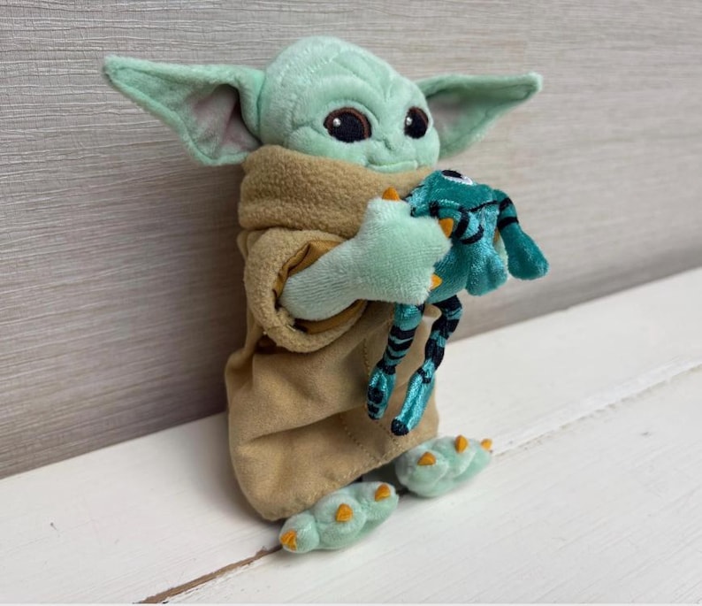 May include: A plush Grogu doll, also known as Baby Yoda, in a tan robe, holding a teal frog toy. The doll has large black eyes, pointed ears, and green skin. The frog toy has black and teal stripes.