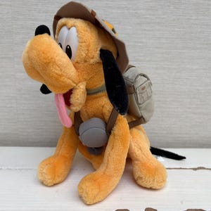 May include: A plush toy of Pluto, the Disney cartoon dog, in a seated position. Pluto is orange with black ears, a pink tongue, and a brown hat and backpack. The backpack is tan and gray, and the hat is brown.