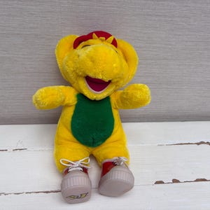 Puede incluir: Un peluche de un personaje de dinosaurio amarillo con una barriga verde, una gorra roja en la cabeza y zapatillas rojas con cordones blancos. El personaje tiene la boca abierta mostrando un interior morado y está de pie.