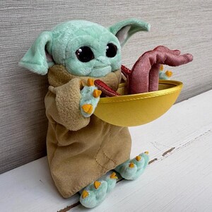 Pode incluir: Um brinquedo de pel&uacute;cia de um personagem de uma popular s&eacute;rie de fic&ccedil;&atilde;o cient&iacute;fica. A figura tem grandes olhos pretos, pele verde e orelhas grandes. Ele est&aacute; vestindo uma t&uacute;nica marrom e segurando uma tigela dourada com um objeto vermelho dentro. Os p&eacute;s e as m&atilde;os do brinquedo s&atilde;o verde claro com detalhes amarelos.