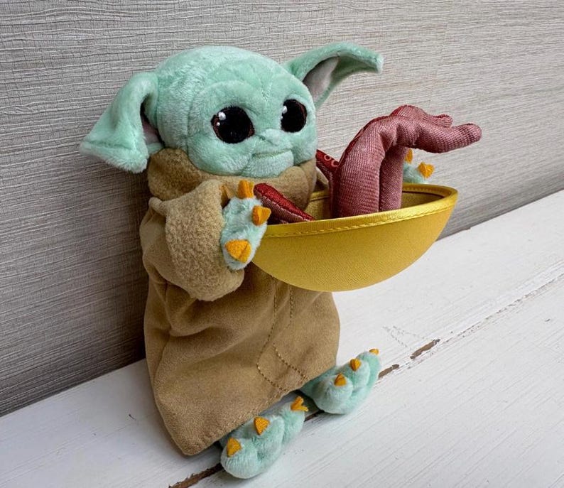 Pode incluir: Um brinquedo de pel&uacute;cia de um personagem de uma popular s&eacute;rie de fic&ccedil;&atilde;o cient&iacute;fica. A figura tem grandes olhos pretos, pele verde e orelhas grandes. Ele est&aacute; vestindo uma t&uacute;nica marrom e segurando uma tigela dourada com um objeto vermelho dentro. Os p&eacute;s e as m&atilde;os do brinquedo s&atilde;o verde claro com detalhes amarelos.
