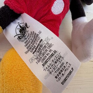 K&ouml;nnte beinhalten: Ein Mickey Mouse Pl&uuml;schtier mit rotem Oberteil, schwarzer Hose, gelben Schuhen und wei&szlig;en Handschuhen. Ein Disney-Etikett ist sichtbar.