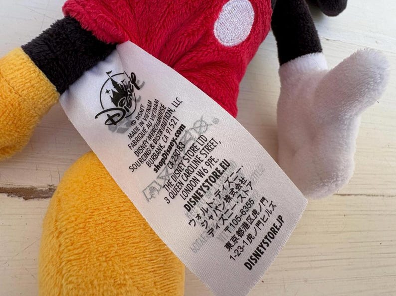 K&ouml;nnte beinhalten: Ein Mickey Mouse Pl&uuml;schtier mit rotem Oberteil, schwarzer Hose, gelben Schuhen und wei&szlig;en Handschuhen. Ein Disney-Etikett ist sichtbar.