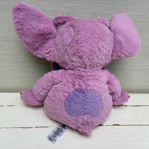 May include: A plush toy in shades of purple, featuring large, floppy ears and a heart-shaped patch on its rear. The toy is made of soft, fuzzy material and has a tag attached.