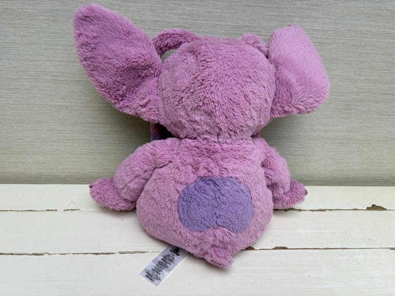 May include: A plush toy in shades of purple, featuring large, floppy ears and a heart-shaped patch on its rear. The toy is made of soft, fuzzy material and has a tag attached.