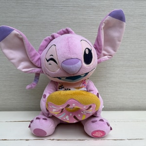 Puede incluir: Un peluche de un personaje alien&iacute;gena morado con orejas grandes, gui&ntilde;ando un ojo y sosteniendo una rosquilla amarilla y rosa. El juguete tiene una cara detallada y est&aacute; sentado erguido. El fondo es gris claro.