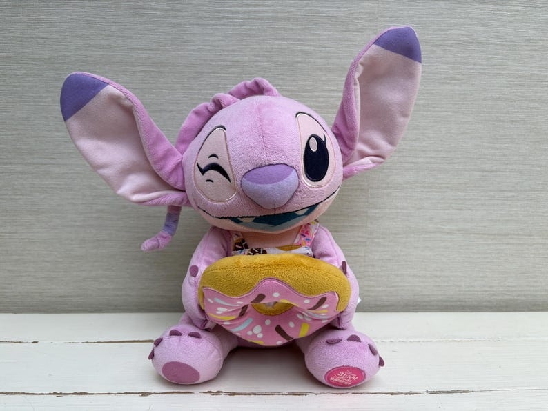 Puede incluir: Un peluche de un personaje alien&iacute;gena morado con orejas grandes, gui&ntilde;ando un ojo y sosteniendo una rosquilla amarilla y rosa. El juguete tiene una cara detallada y est&aacute; sentado erguido. El fondo es gris claro.