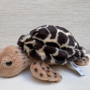 May include: A plush turtle toy with a brown and cream shell pattern. The turtle's head and flippers are tan with black spots. The toy has a tag attached. This soft toy is suitable for children.