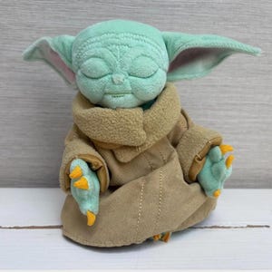 May include: A plush toy of a character with large ears, green skin, and closed eyes. The character is wearing a tan robe with a brown collar and has three-fingered hands with orange claws. The toy is sitting on a white surface.