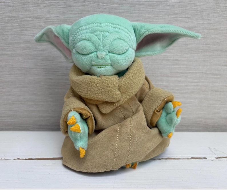 May include: A plush toy of a character with large ears, green skin, and closed eyes. The character is wearing a tan robe with a brown collar and has three-fingered hands with orange claws. The toy is sitting on a white surface.