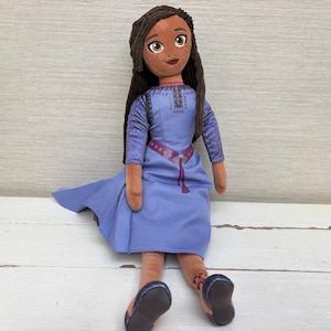 May include: A plush doll with long dark brown hair, brown eyes, and freckles, wearing a light purple dress with a printed pattern on the bodice and waist. The doll has tan skin, long sleeves, and dark blue shoes with a small decorative detail on one ankle.