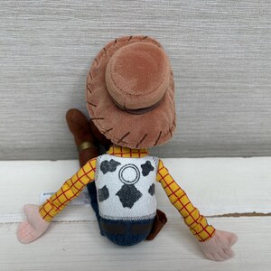 May include: A plush toy of a cowboy character, viewed from the back. The toy has a brown hat, a yellow plaid shirt, and a white vest with a black cowhide pattern. The arms and hands are pink, and the jeans are blue.