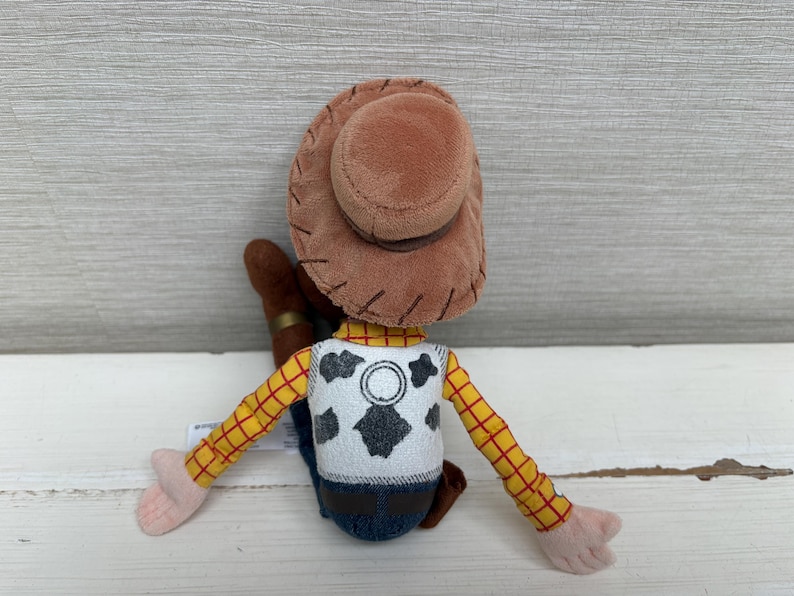 May include: A plush toy of a cowboy character, viewed from the back. The toy has a brown hat, a yellow plaid shirt, and a white vest with a black cowhide pattern. The arms and hands are pink, and the jeans are blue.