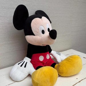 May include: A plush Mickey Mouse doll, featuring a black head and ears, a tan face, and a black nose. The doll wears a black shirt, red shorts with white buttons, white gloves, and large yellow shoes. The toy is sitting on a white surface.