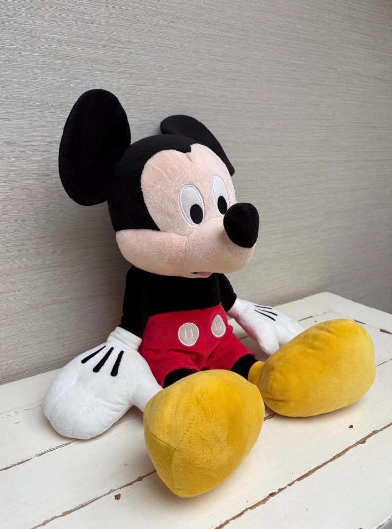 May include: A plush Mickey Mouse doll, featuring a black head and ears, a tan face, and a black nose. The doll wears a black shirt, red shorts with white buttons, white gloves, and large yellow shoes. The toy is sitting on a white surface.