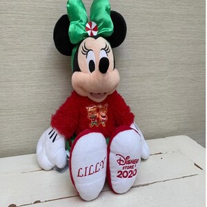 May include: A plush Minnie Mouse doll wearing a red sweater and a green bow. The doll has black ears, white gloves, and red and white shoes. The word "LILLY" is embroidered on one shoe, and "Disney Store 2020" is on the other.