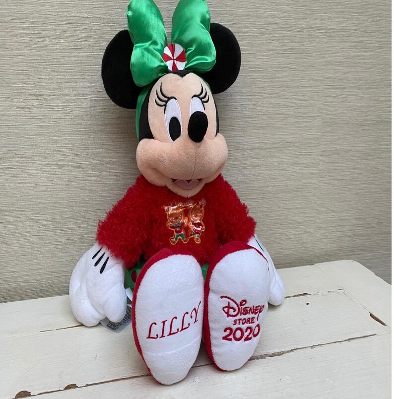 May include: A plush Minnie Mouse doll wearing a red sweater and a green bow. The doll has black ears, white gloves, and red and white shoes. The word "LILLY" is embroidered on one shoe, and "Disney Store 2020" is on the other.