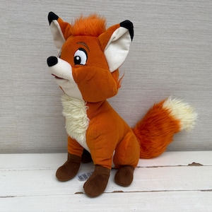 May include: A plush fox toy featuring vibrant orange fur, a cream-coloured chest, and dark brown paws. The fox has large, expressive eyes, black-tipped ears, and a bushy tail with a cream-coloured tip. It is designed in a seated position.