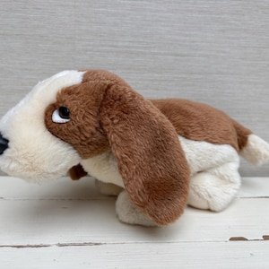 Può includere: Un peluche a forma di cane basset hound marrone, bianco e nero. L'animale di peluche ha lunghe orecchie flosce, un petto bianco e una schiena marrone. Il giocattolo è appoggiato su una superficie bianca.